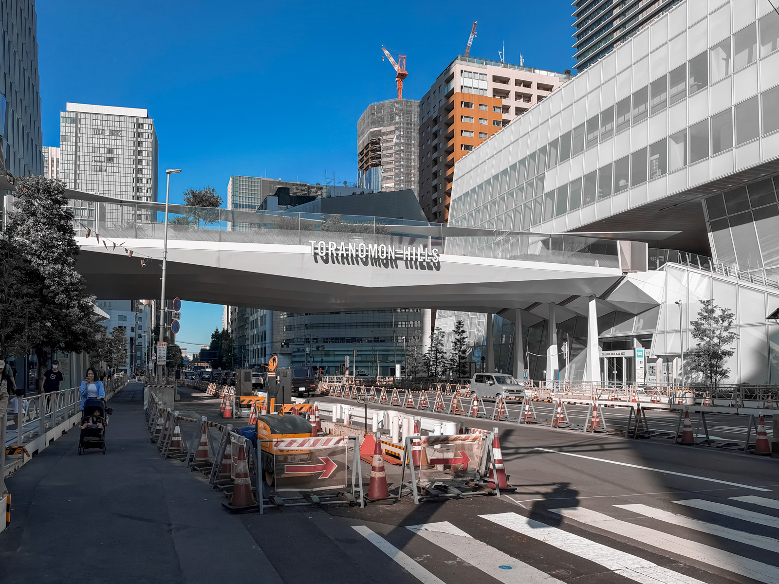 「TORANOMON HILLS」の白い文字が刻まれた歩道橋デッキ。Hotel Toranomon Hillsの看板。奥に建設中のクレーンと工事中の道路