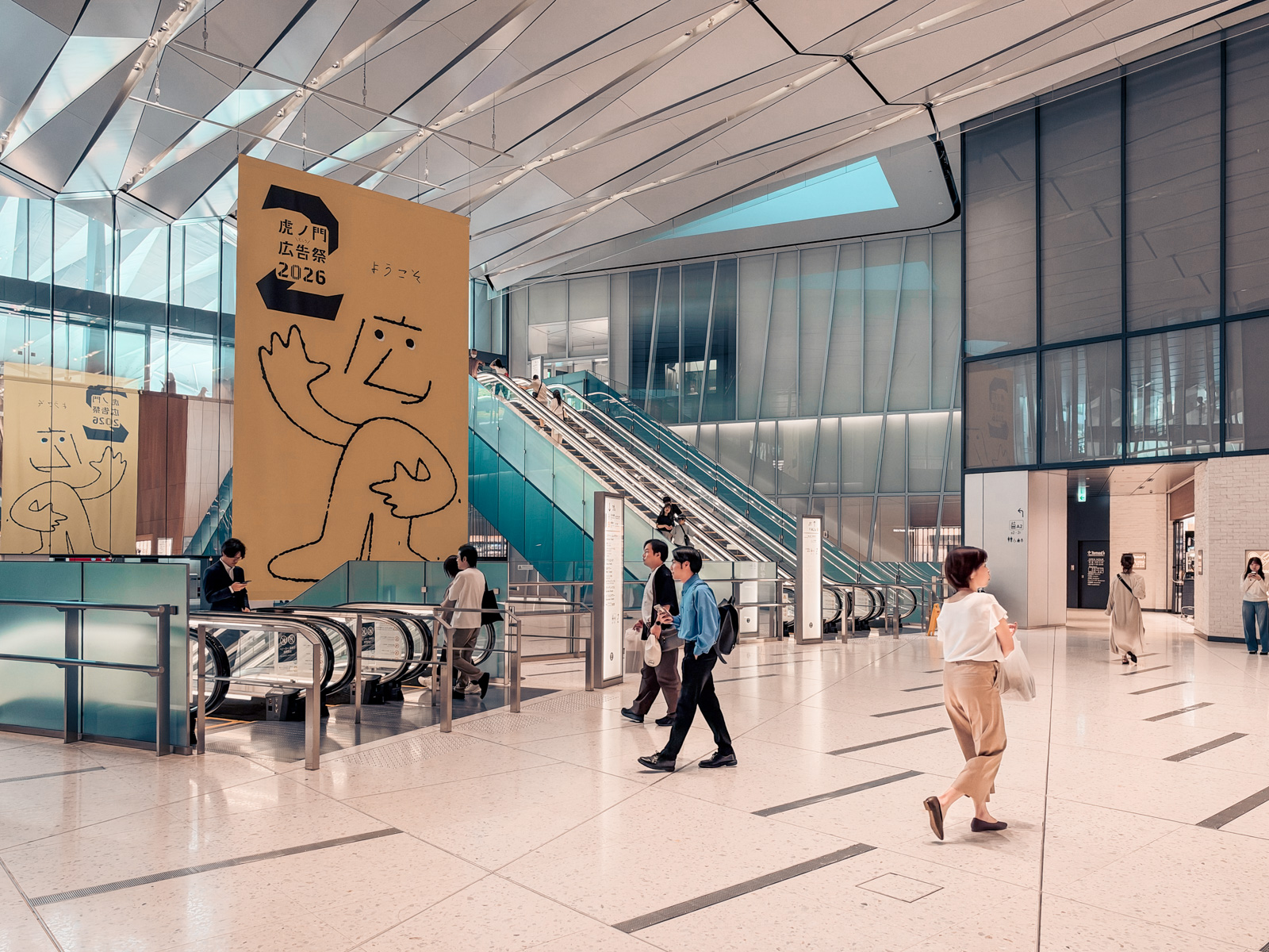 「虎ノ門広告祭2026」の大型バナー。黄色い背景にキャラクターの線画。その前を人が通り過ぎる。青いエスカレーターが奥に続く