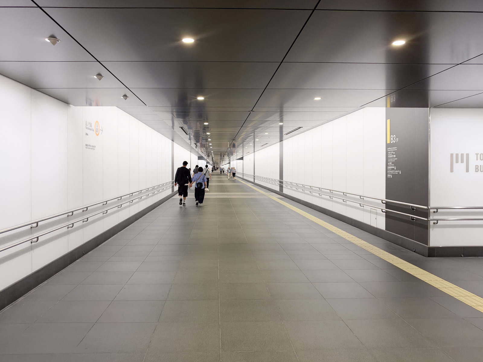 地下連絡通路B3。長い直線の廊下。白い壁、灰色の床、定間隔の照明。数人が歩いている。右側に「TO BU」の文字が見える案内板