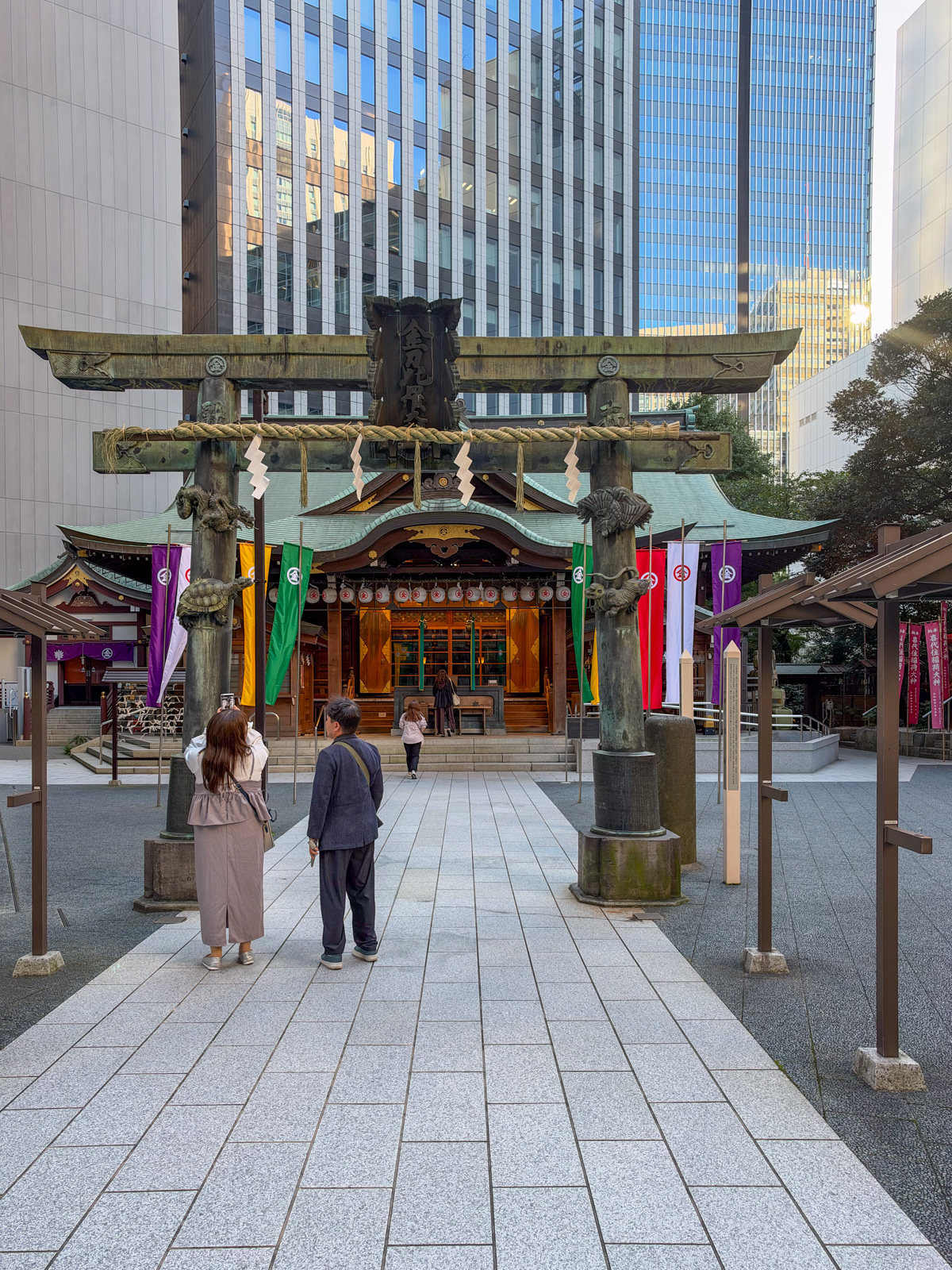 金刀比羅宮 拝殿正面。大きな銅製鳥居、カラフルな幟(紫・黄・緑・赤・白)、社殿。参拝者が二人。背後にタワービル群と空
