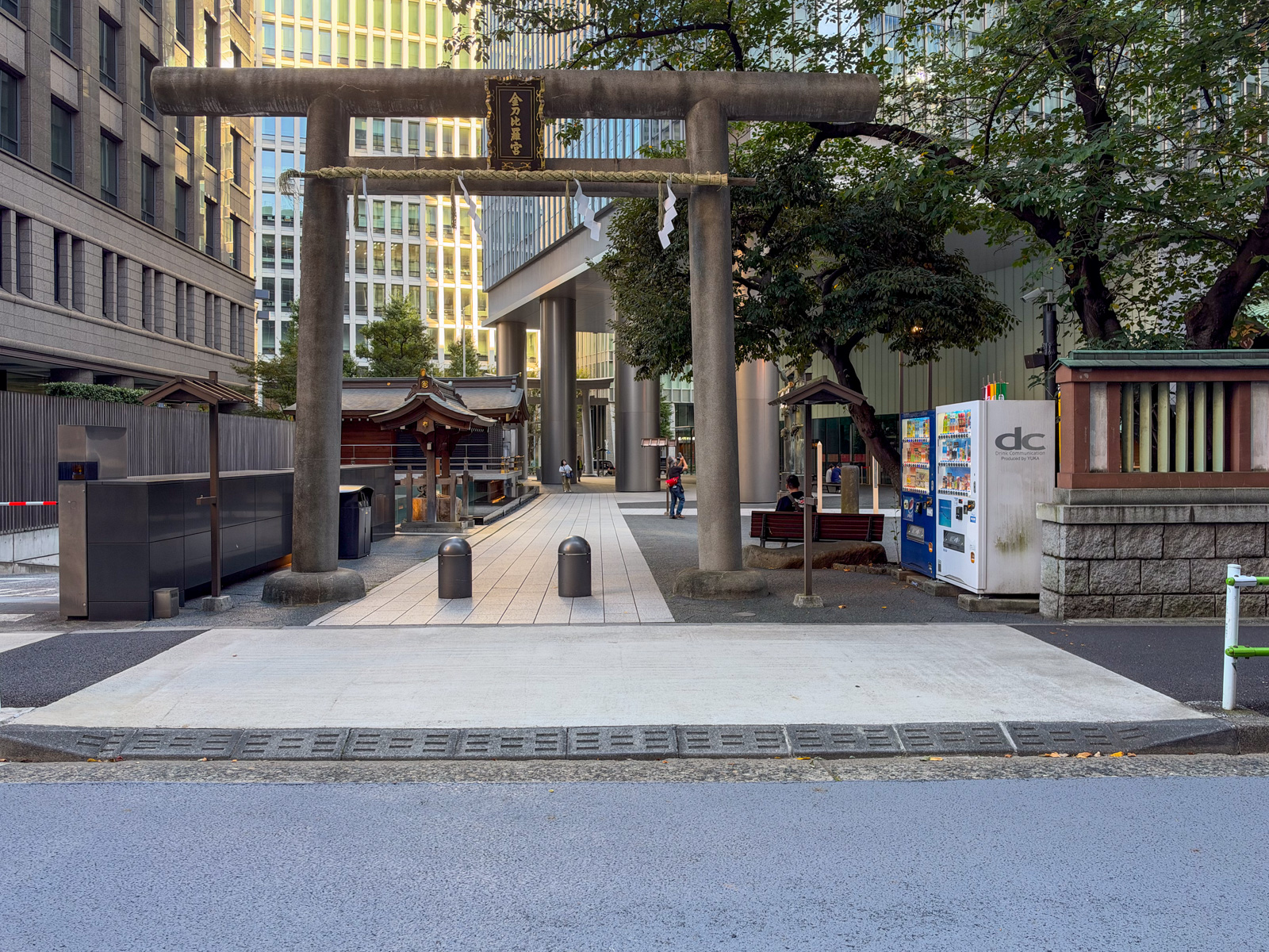 街路の鳥居。「金刀比羅宮」の扁額が掲げられた石鳥居。鳥居越しに境内と社殿が見える。背後に自販機とタワービル群